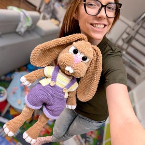 May include: A handmade brown crochet bunny doll with large ears, black eyes, and a pink nose. The bunny wears purple overalls with yellow and white striped shirt. The doll is soft and plush, perfect for a child's toy or nursery decor.