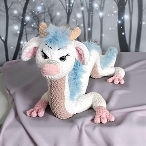May include: A handmade crochet dragon toy in white, light blue, and pink. The dragon has a long, serpentine body with four legs, a pink snout, and a fluffy blue mane. The dragon's horns are tan, and its eyes are black.