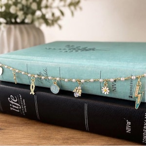 May include: A gold-tone chain bracelet featuring small white faux pearls and multiple charms, including a heart, a cross, a fish symbol, a round blue WWJD pendant, an angel, a white daisy, and a teal lightning bolt with gold stars.