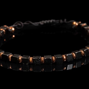 May include: A bracelet featuring a repeating pattern of dark metallic cube-shaped beads and smaller, smooth copper-coloured circular spacer beads. The bracelet is arranged in a circular shape and includes a dark, twisted wire clasp mechanism.