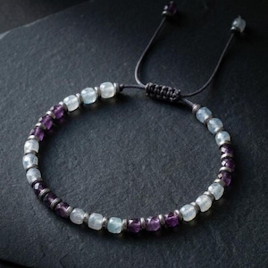May include: A beaded bracelet featuring alternating faceted purple amethyst and translucent light blue moonstone beads separated by small silver-toned metal spacers. The bracelet includes a black cord with an adjustable sliding knot closure.