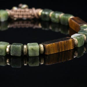Op de afbeelding: Een close-up van een kralenarmband met afwisselend groene vierkante kralen en bruine rechthoekige kralen. De armband is geregen op een beige koord en heeft goudkleurige metalen afstandhouders. De armband wordt gepresenteerd op een reflecterend zwart oppervlak.