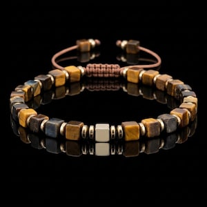 Può includere: Un bracciale con perline cubiche in occhio di tigre nei toni del marrone e del blu, alternate a piccoli distanziatori metallici. Un singolo cubo centrale in metallo dorato liscio e lucido funge da elemento principale, con chiusura a cordino marrone.