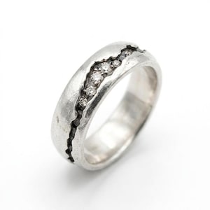 May include: A silver ring with a textured surface and a crack-like design filled with small, sparkling stones. The ring has a band width of approximately 0.25 inches.