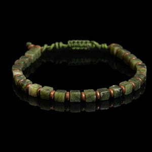 May include: A handmade bracelet featuring square green gemstone beads and copper-colored spacers. The bracelet is strung on a green cord and is displayed against a black background. This piece of jewelry is suitable for everyday wear.