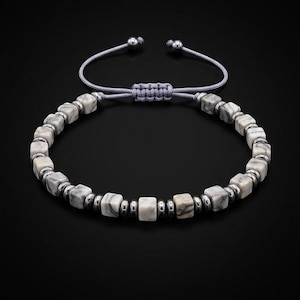 Op de afbeelding: Een armband met afwisselende kubusvormige kralen met een grijs en wit marmerpatroon en kleine, gepolijste zilverkleurige metalen tussenkralen. De armband heeft een grijs koord met een verstelbaar schuifknoopsluitingsmechanisme.