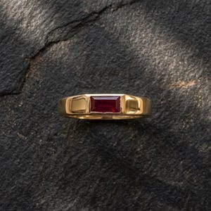 May include: A gold ring with a rectangular, deep red gemstone set in the center. The ring has a simple, polished band and a classic design. The background is a dark, textured surface, providing contrast to the ring's shine.