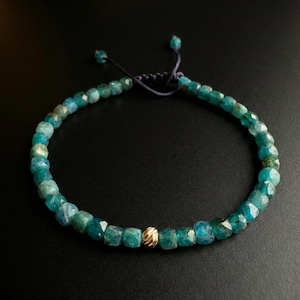 Könnte beinhalten: Ein Armband mit facettierten, türkisblauen Edelsteinen und einer kleinen, strukturierten Goldperle. Das Armband ist an einer dunkelblauen Kordel mit verstellbaren Knoten befestigt. Der Schmuck wird vor einem schwarzen Hintergrund präsentiert.