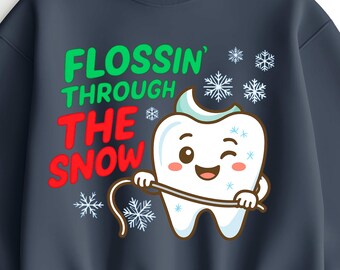 Może przedstawiać: Granatowa bluza z napisem "FLOSSIN' THROUGH THE SNOW" w kolorze zielonym i czerwonym. Kreskówkowy ząb używa nici dentystycznej, otoczony płatkami śniegu. Zabawny design dla dentystów lub miłośników humoru dentystycznego.
