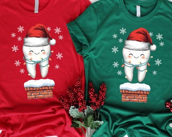 May include: Two short-sleeved t-shirts, one red and one green, each featuring a cartoon tooth wearing a Santa hat and wrapped in Christmas lights. The tooth is smiling and standing on a brick chimney with snowflakes around it. Festive holiday apparel.