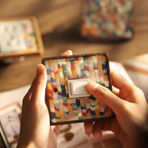 May include: A decorative light switch with a colourful, abstract pattern. The switch plate has a wooden frame and a white toggle switch. The design features a mix of red, yellow, blue, and green hues. The switch is being held.