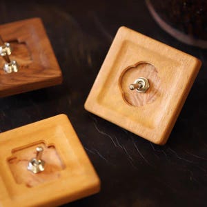 May include: Three wooden light switch plates are shown. The plates are square, with a light wood finish and a silver toggle switch. One plate has a decorative floral-shaped cutout around the switch. The switches are set against a dark background.