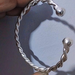 Könnte beinhalten: Ein silberfarbenes Armreif-Armband mit einem gedrehten Seildesign. Die Enden des Armbands haben eine abgerundete, kugelförmige Form. Das Armband wird von einer Hand vor einem hellblauen Hintergrund gehalten.