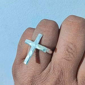 以下が含まれることがあります： 十字架の形をした銀色の指輪。 指輪はハンマーで叩いたような質感で、指に装着されています。 背景は水色です。