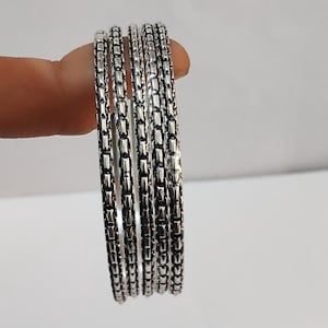 May include: Five silver chain link bracelets are stacked together. Each bracelet is made of small, rectangular links, creating a textured surface. The bracelets are displayed together, highlighting their design and shine. The background is a soft, neutral colour.