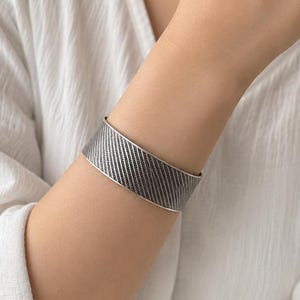 May include: A wide silver cuff bracelet featuring a textured, diagonal cross-hatch pattern across its entire surface. The metal band has a polished, smooth edge and a consistent width, wrapping around the wrist with a dark, oxidized finish.