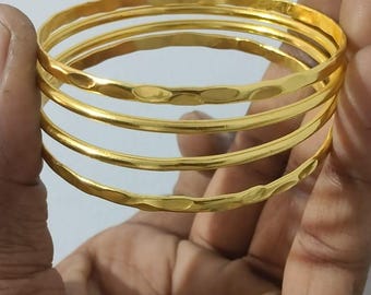 以下が含まれることがあります： 金色のバングル5本セット。バングルはハンマー加工されたテクスチャです。白い背景に、手で持たれています。シンプルでエレガントなアクセサリーです。