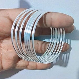 以下が含まれることがあります： 銀色のバングルが積み重ねられています。細くて丸いブレスレットが重ねられ、レイヤード効果を生み出しています。バングルは、無地の背景に対して、手で持たれています。