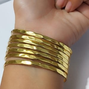 Può includere: Una pila di braccialetti color oro con una texture martellata adorna un polso. I braccialetti sono impilati, creando un accessorio audace e accattivante. I gioielli sono realizzati in metallo lucido e riflettente.