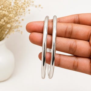 May include: Two smooth, polished silver-toned bangle bracelets with a rounded, circular profile. The bangles feature a reflective, high-shine finish and a simple, continuous band design without any additional embellishments or patterns.