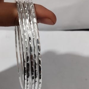 May include: A stack of silver bangles with a hammered texture. The bangles are round and have a shiny, reflective surface. The image is a close-up shot, highlighting the texture and design of the jewelry.