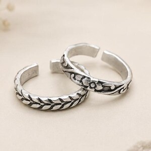 Puede incluir: Dos anillos abiertos de tono plateado con detalles oxidados. Un anillo presenta un patrón repetitivo de hojas o trigo, mientras que el otro muestra un motivo floral con pequeños pétalos y enredaderas. Ambos anillos tienen una abertura.