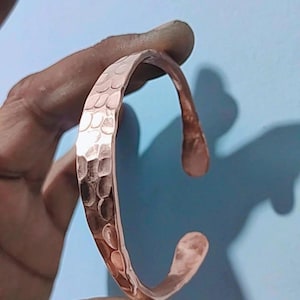 May include: A copper cuff bracelet with a hammered texture. The open-ended bracelet has a curved shape and is held against a light blue background. The bracelet's surface reflects light, highlighting its metallic sheen.