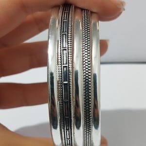 May include: A stack of silver bangles with detailed designs. The bangles have a polished finish, with one featuring a rectangular pattern and another with a dotted texture. The jewellery is held by a hand.