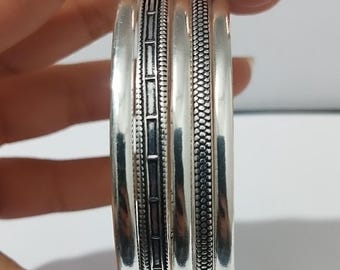 May include: A stack of silver bangles with detailed designs. The bangles have a polished finish, with one featuring a rectangular pattern and another with a dotted texture. The jewelry is held by a hand.