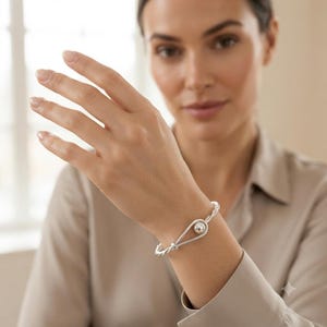 Può includere: Un bracciale color argento caratterizzato da una fascia in metallo intrecciato che forma un occhiello a goccia al centro. Una sfera d'argento liscia e lucida è posizionata all'interno dell'occhiello, fissata dalla struttura intrecciata.