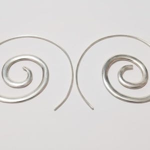 Puede incluir: Un par de pendientes plateados en forma de espiral. Cada pendiente presenta un alambre curvado que forma una espiral con un centro redondeado. Se muestran sobre un fondo blanco liso.