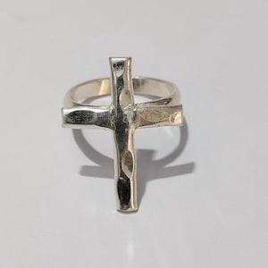 May include: A silver-colored ring featuring a textured cross design. The cross is the primary focus, with a hammered metal appearance. The ring band is smooth and simple, complementing the cross's detailed texture. The ring is set against a white background.
