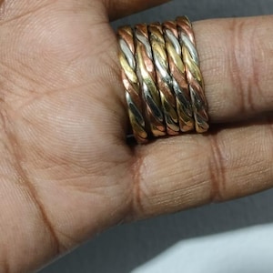 Pode incluir: Um conjunto de seis anéis de metal torcido em várias cores, incluindo ouro, prata e cobre. Os anéis são empilhados, mostrando um design trançado. Os anéis estão na mão de uma pessoa.
