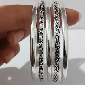 May include: A set of silver bangles, some with floral and leaf patterns. The bangles are smooth and shiny, with detailed designs. The image shows the bangles held in a hand, showcasing the jewelry's design and texture.