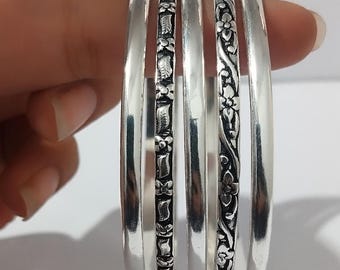May include: A set of silver bangles, some with floral and leaf patterns. The bangles are smooth and shiny, with detailed designs. The image shows the bangles held in a hand, showcasing the jewelry's design and texture.