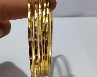 以下が含まれることがあります： テクスチャ加工された表面を持つ、6つのゴールドバングルのセット。指で持たれています。金は光を反射し、白い背景にハイライトと影を作り出します。各バングルは約6cmの直径です。