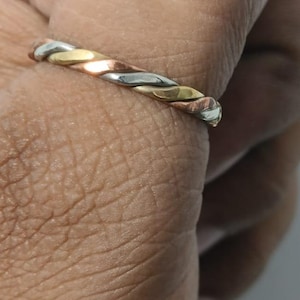 Può includere: Un anello intrecciato con una combinazione di metalli argentati, dorati e ramati. L'anello è indossato su un dito, mettendo in risalto il design intrecciato e le tonalità metalliche. Un gioiello semplice ma elegante.