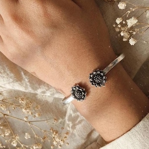 Könnte beinhalten: Silbernes Armreif-Armband mit zwei detaillierten Sonnenblumen-Designs. Das offene Armband ermöglicht eine einfache Anpassung. Die Sonnenblumen sind aufwendig gearbeitet und verleihen dem Schmuck einen Hauch von naturnaher Eleganz.
