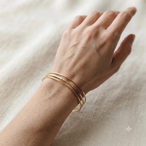 Puede incluir: Un conjunto de tres brazaletes finos de oro pulido que se llevan en la muñeca. Las pulseras son circulares, lisas y metálicas, con una superficie reflectante que capta la luz. Cada brazalete es idéntico en tamaño y forma, creando un efecto apilado.