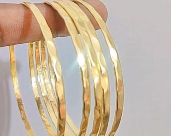 May include: A collection of six gold-coloured bangles with a hammered texture. The bracelets are round and have a shiny finish. The bangles are held together, showcasing their design and colour.