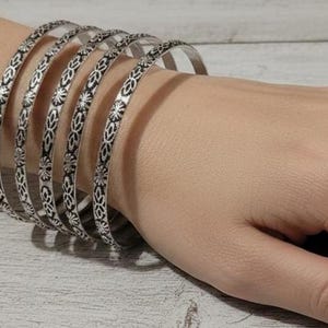 Könnte beinhalten: Eine Reihe silberner Armreifen mit aufwendigen floralen Mustern. Die Armreifen sind an einem Handgelenk gestapelt und zeigen ihr detailliertes Design. Die Armbänder bestehen aus glänzendem Metall und verleihen dem Gesamtbild einen Hauch von Eleganz.