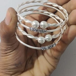 Può includere: Una collezione di braccialetti d'argento. Alcuni braccialetti hanno un design a corda intrecciata, mentre altri presentano perline decorative e dettagli a motivi. I braccialetti sono esposti in una mano.