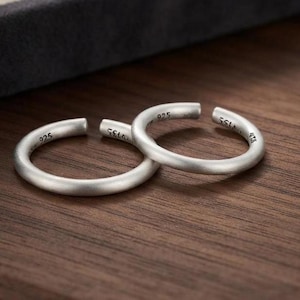 Könnte beinhalten: Zwei silberfarbene offene Ringe mit matter Oberfläche. Jeder Ring trägt die Prägung 925 auf der Innenseite, was auf die Metallzusammensetzung hinweist. Die Ringe haben ein schlichtes, kreisförmiges Banddesign mit einer kleinen Öffnung.