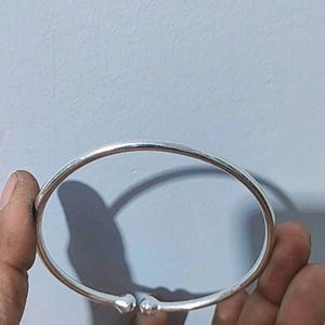 Könnte beinhalten: Ein silberfarbenes Armreif-Armband mit einem einfachen, abgerundeten Design. Das Armband hat eine glatte, polierte Oberfläche und verfügt über zwei kleine, abgerundete dekorative Elemente an der Öffnung. Das Armband wird in einer Hand vor einem hellblauen Hintergrund gehalten.