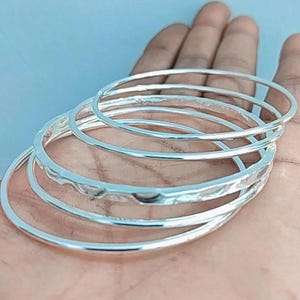 May include: Several silver bangles, some with a hammered texture, are shown. The thin, circular bangles reflect light. The image shows the bangles resting on a hand, against a light blue background.