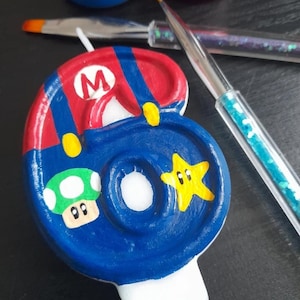 Puede incluir: Una vela n&uacute;mero seis pintada a mano, con un dise&ntilde;o azul y rojo inspirado en el personaje Mario. La vela incluye un hongo verde y una estrella amarilla. Pinceles y botes de pintura en el fondo.