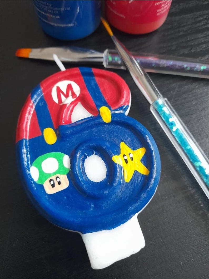 Puede incluir: Una vela n&uacute;mero seis pintada a mano, con un dise&ntilde;o azul y rojo inspirado en el personaje Mario. La vela incluye un hongo verde y una estrella amarilla. Pinceles y botes de pintura en el fondo.