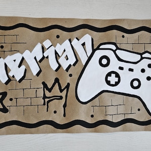 May include: A rectangular brown paper sign featuring the name Kierian in white graffiti-style lettering, a white video game controller illustration, a black crown, and two black stars, all set against a hand-drawn black brick wall pattern.
