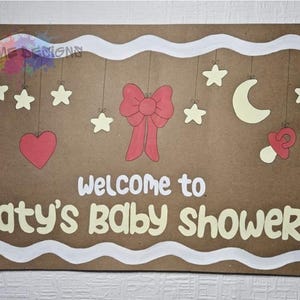 Può includere: Un cartello marrone con bordo bianco e la scritta "welcome to katy's baby shower". Il cartello presenta stelle sospese, un cuore, un fiocco, una luna e un ciuccio. Il design è nei toni del rosso, crema e bianco.