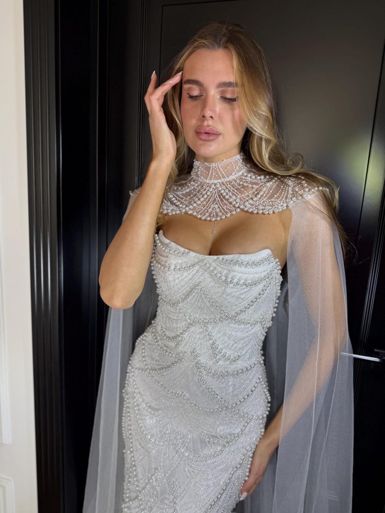Strapless mermaid wedding dress with pearl embellishments, sculpted corset bodice and elegant sheer bridal cape in ivory lace.